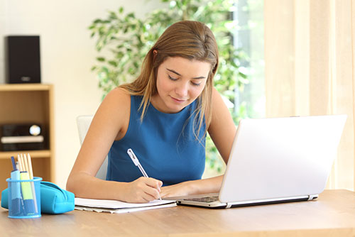 young female writing at desk with laptop